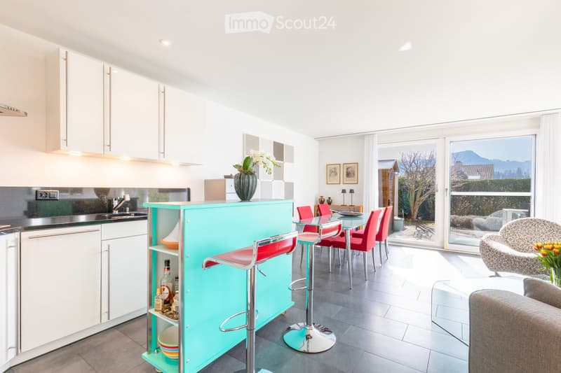 Modern kitchen and dining area with white cabinets, a turquoise bar counter, red stools, a glass dining table with red chairs, a grey sofa, and a vase with flowers on the counter.