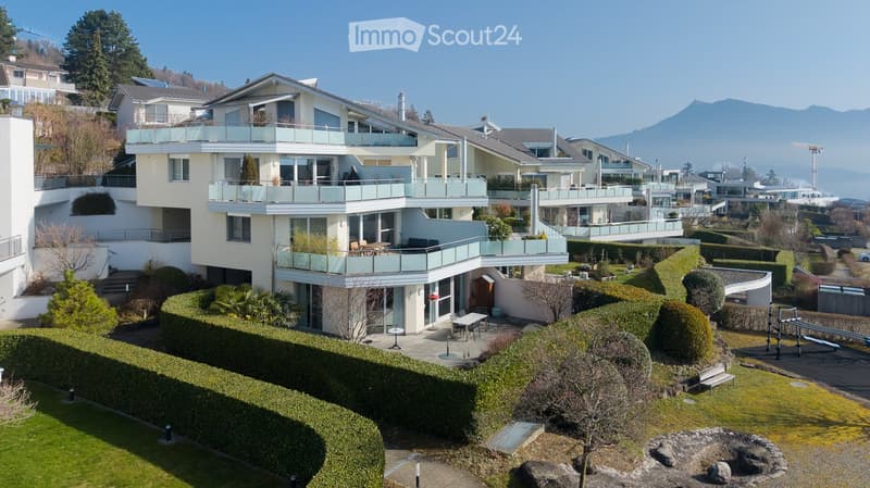 Modern multi-story residential building with balconies, surrounded by landscaped gardens and trees, overlooking a scenic mountain view.