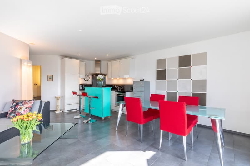 Modern open-plan living and dining area with a grey sofa, glass coffee table, glass dining table with red chairs, turquoise bar cabinet, and kitchen area with white cabinets and stainless steel appliances.
