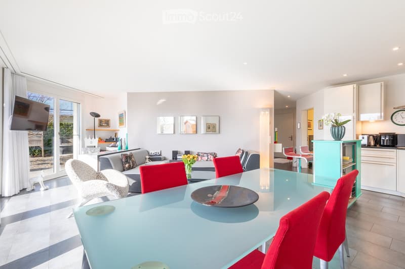 A modern, open-plan living and dining area with a glass dining table, red chairs, a grey sofa, a television, and a kitchen area with cabinets and appliances.
