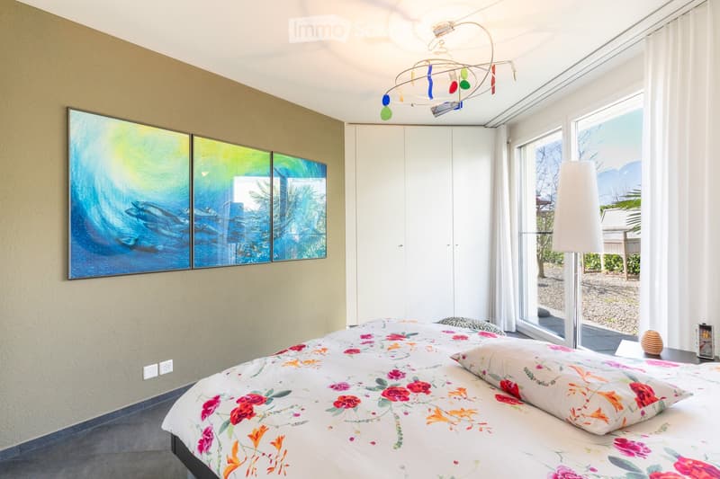 Modern bedroom with a large bed, floral duvet, white wardrobe, floor lamp, and large windows with a scenic view.