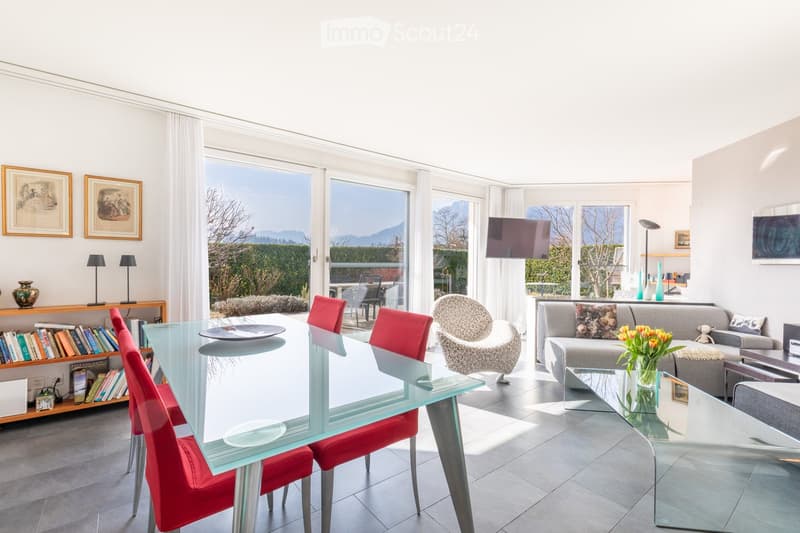 Bright living and dining area with large windows and a view, featuring a grey sofa, glass coffee table, glass dining table with red chairs, leather armchair, bookshelf, and decorative vases.