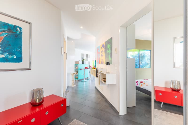 Modern hallway with white walls, grey tiled floor, and several rooms visible including a kitchen area with colorful stools, a bedroom with a bed, and a living area with a red cabinet and decorative items.