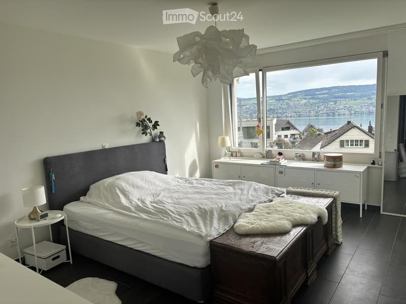 Modern bedroom with a large bed, nightstand, lamp, and a scenic view through large windows.