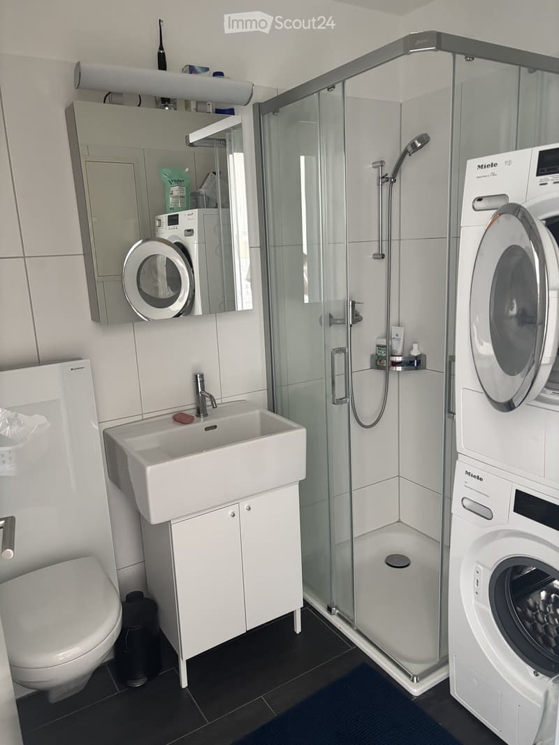 Modern bathroom with white tiles, a glass-enclosed shower, a sink with a mirror, a toilet, and a washing machine.