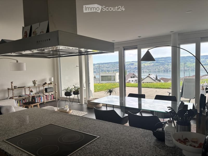 Open-plan living and dining area with modern furniture, large windows offering a scenic view, and a kitchen area with an induction stove and countertop.