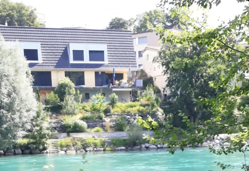 A modern two-story house with a sloped roof, large windows, and a balcony overlooking a serene body of water, surrounded by lush greenery and trees.