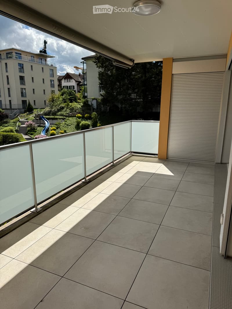 Modern balcony with grey tiled floor, glass railing, and view of a landscaped garden and neighboring buildings.