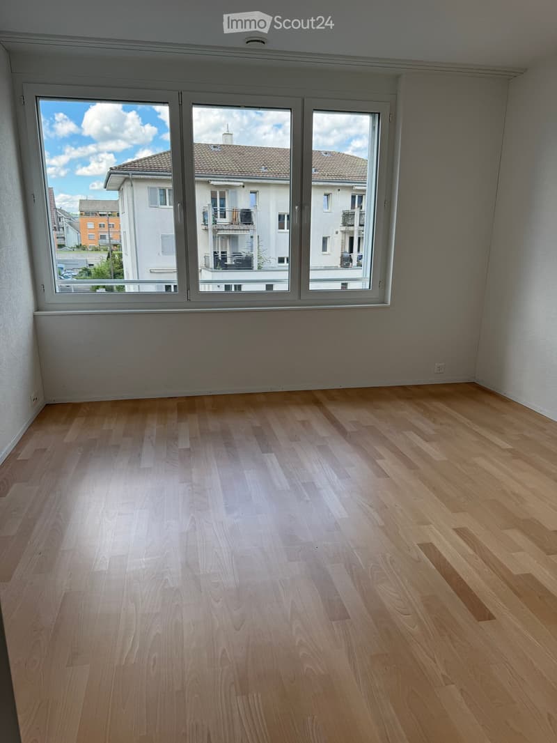 Empty room with wooden flooring and large windows showing a view of neighboring buildings and a clear sky.