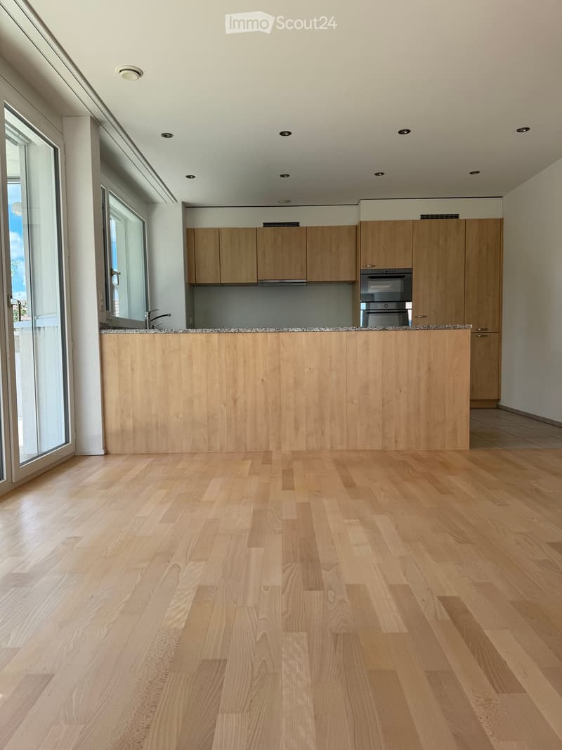 Empty kitchen with light wooden cabinets, a granite countertop, and a wooden floor. Large windows and a sliding door provide natural light.