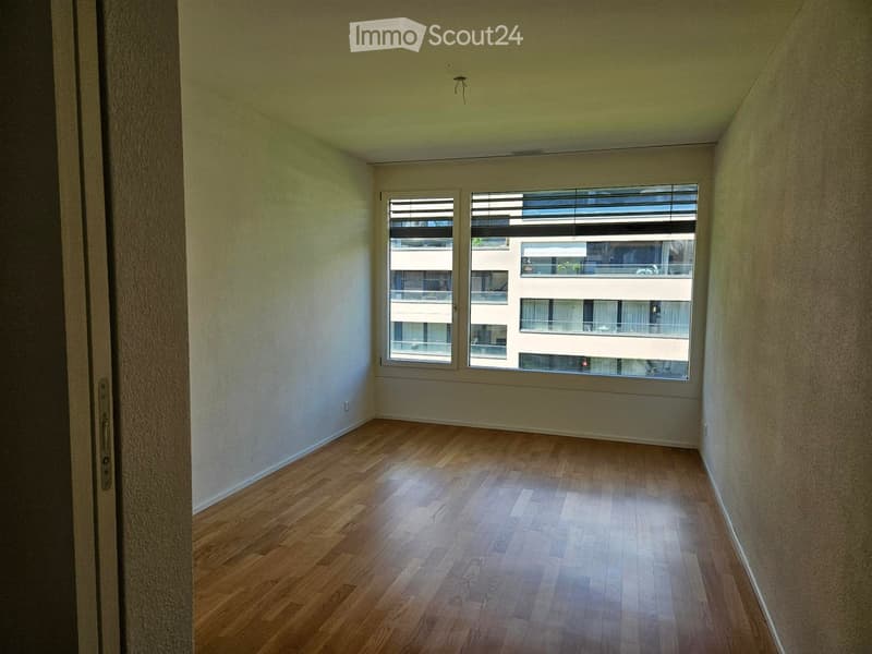 Empty room with wooden flooring, white walls, and large windows showing a view of neighboring buildings.