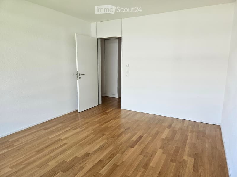 Empty room with light wooden flooring and white walls, featuring an open door leading to another space.