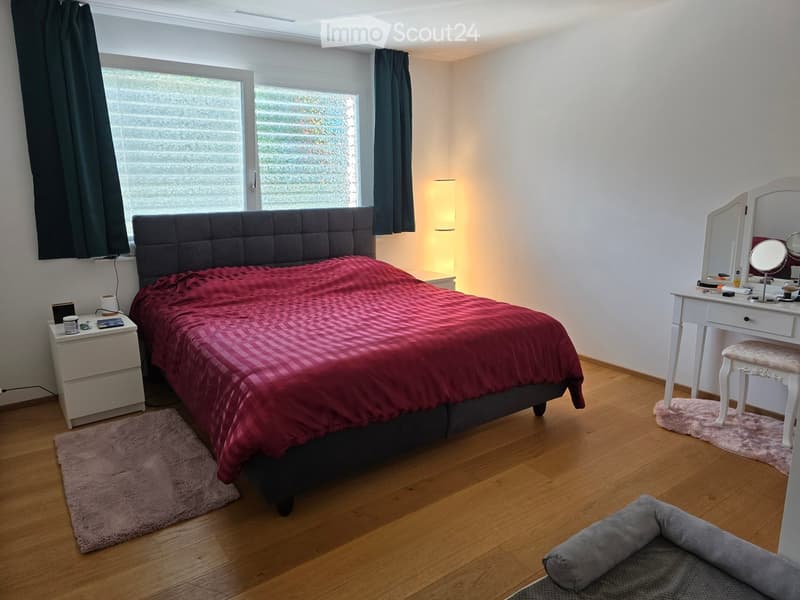 Cozy bedroom with wooden flooring, large window with dark curtains, grey upholstered headboard, red checkered duvet, nightstand with lamp, vanity with mirror and stool, and a pet bed.