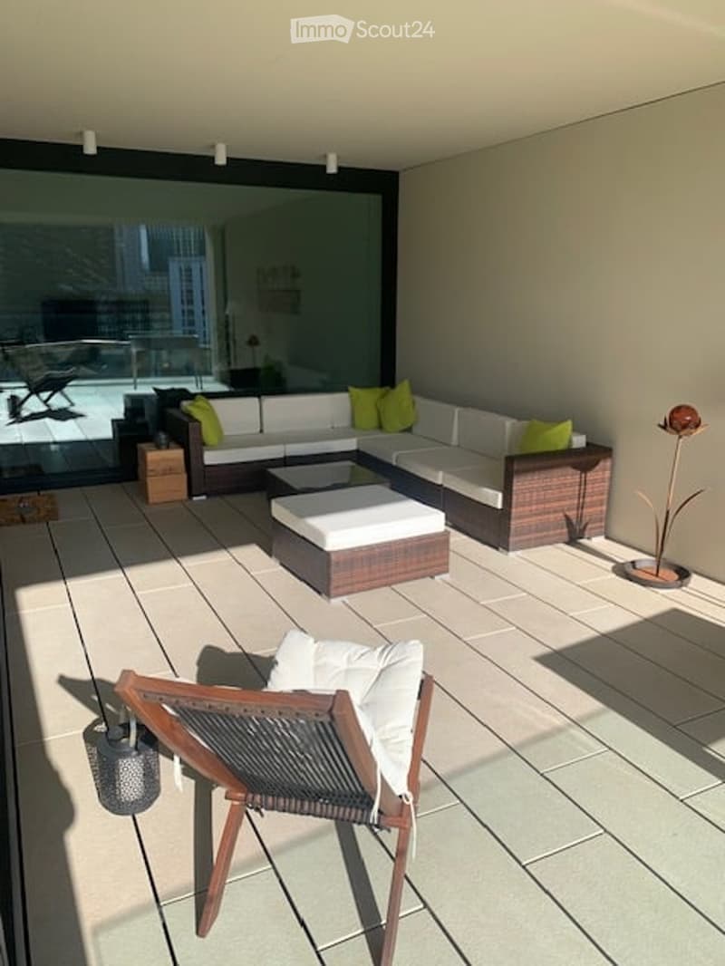 Modern balcony with large glass windows, white sectional sofa, green cushions, wooden coffee table, and a single wooden chair.