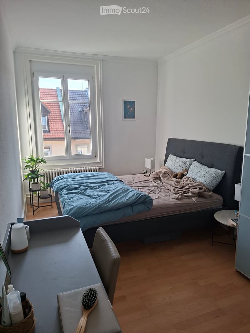 A cozy bedroom with a neatly made bed, a nightstand, a small table with a plant, and a chair near the window. The room features wooden flooring and white walls.