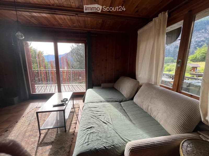 Cozy living room with wooden walls and ceiling, large windows offering a scenic mountain view, a grey sofa, and a coffee table with remote controls.