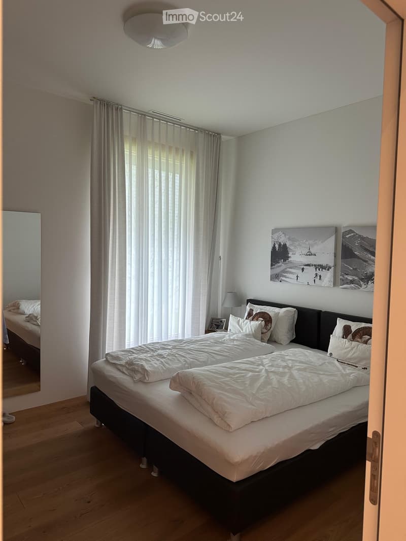 A well-lit bedroom with a large double bed, white bedding, and decorative pillows. The room features wooden flooring, large windows with vertical blinds, a wall-mounted lamp, and artwork depicting a snowy mountain scene.