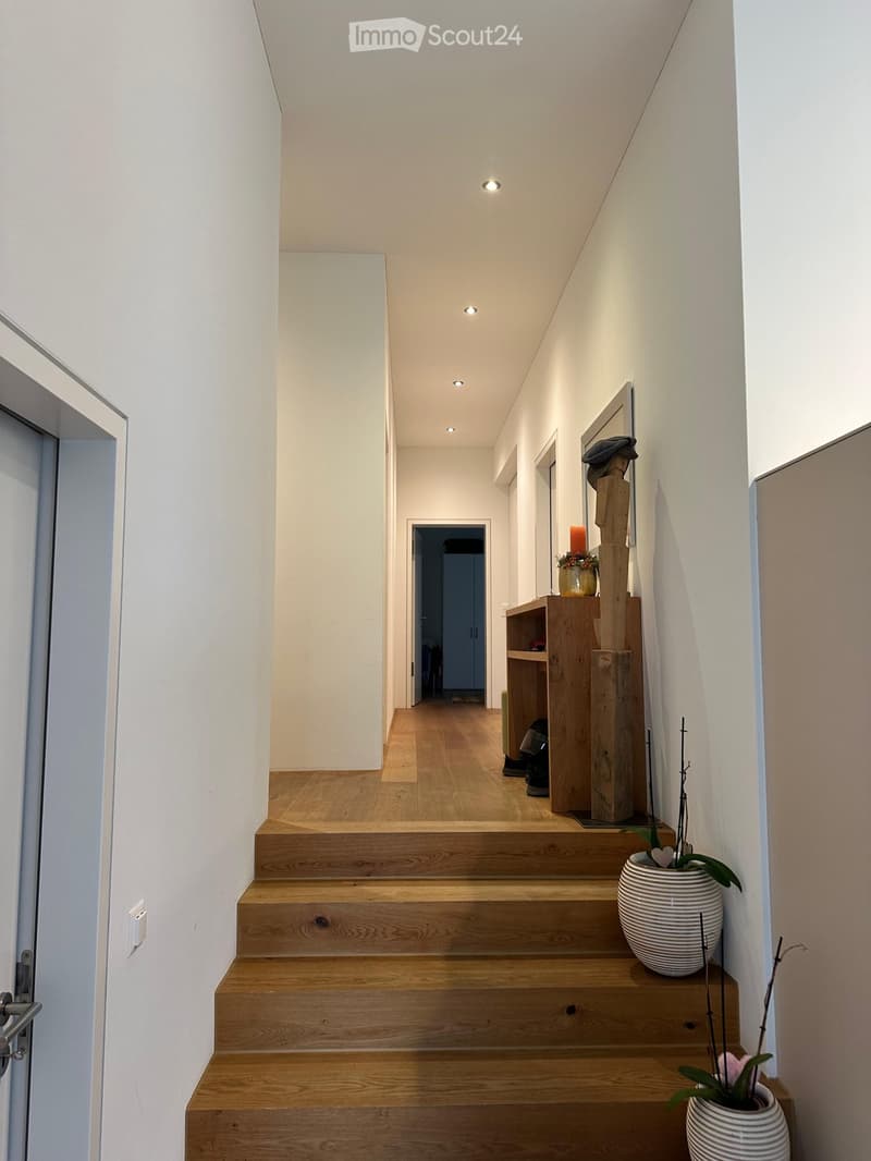 Modern hallway with wooden stairs, white walls, recessed lighting, and decorative items including a vase with flowers and a wooden shelf with shoes and a candle.