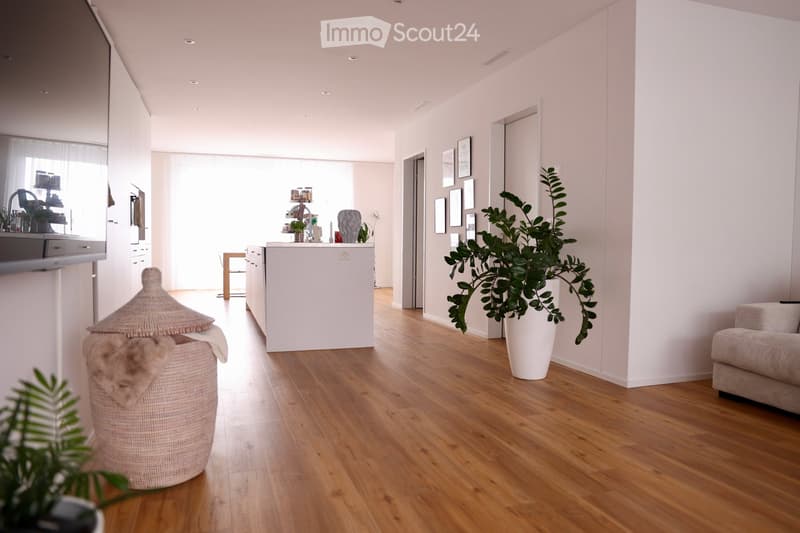 Bright, open living space with light wooden flooring, white walls, and modern decor. Features a sofa, coffee table, kitchen island, framed pictures, and potted plants.