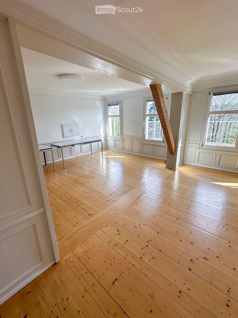 Empty room with light wooden flooring, white walls, and large windows allowing natural light. The room features a modern, minimalist design with a visible structural wooden beam and a small table near the entrance.