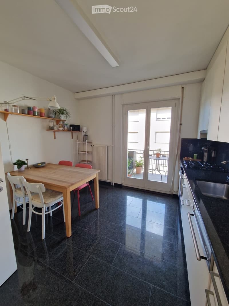 Bright, open-plan kitchen-dining area with dark tiled flooring, white walls, and a large sliding glass door leading to a balcony. The space features a wooden dining table with four chairs, a kitchen counter with a sink and faucet, built-in cabinets, a she
