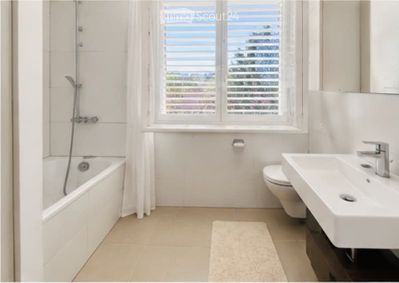 Modern bathroom with a bathtub, toilet, sink, and a window with blinds. The space features white tiles and a light-colored floor.