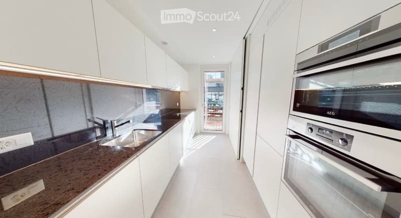 Modern kitchen with white cabinets, dark granite countertops, and stainless steel appliances including a sink and oven. The space is bright with natural light coming through a glass door leading to a balcony.