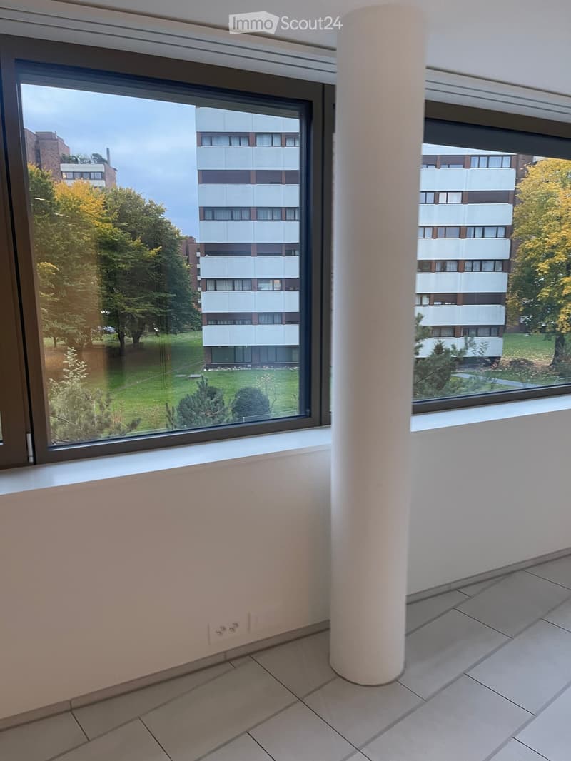 Empty room with tiled floor, light-colored walls, and large windows showing a view of a residential area with trees and buildings.