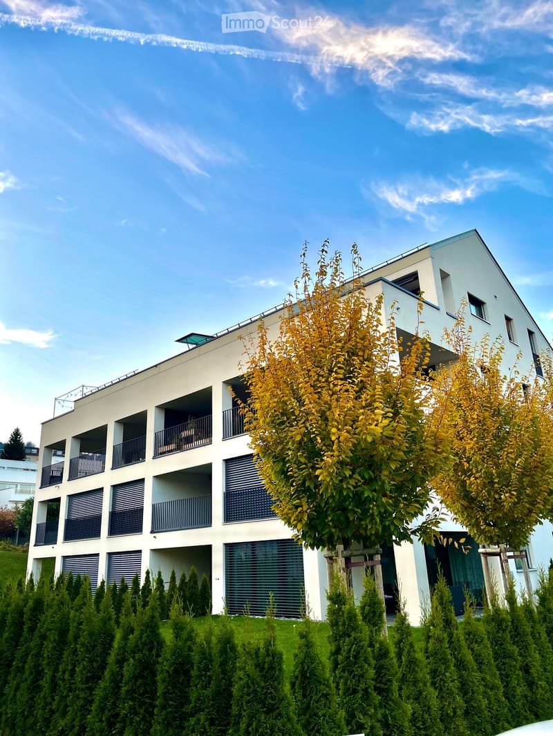 Modern multi-story building with white facade, multiple balconies, and contemporary design, surrounded by neatly trimmed hedges and trees.