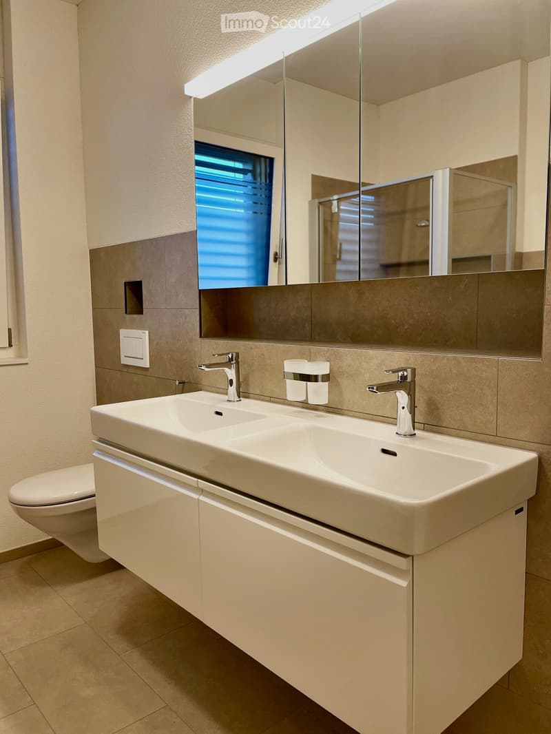 Modern bathroom with a double sink, sleek faucets, a large mirror, and a toilet. The sink has two faucets and two cups placed on it. The walls are tiled in a neutral color scheme, and the floor is tiled as well.