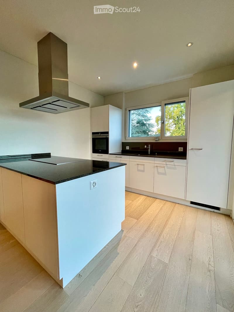 Modern kitchen with white cabinets, black countertops, and a central island. Features include a stainless steel range hood, built-in oven, and a large window overlooking greenery.