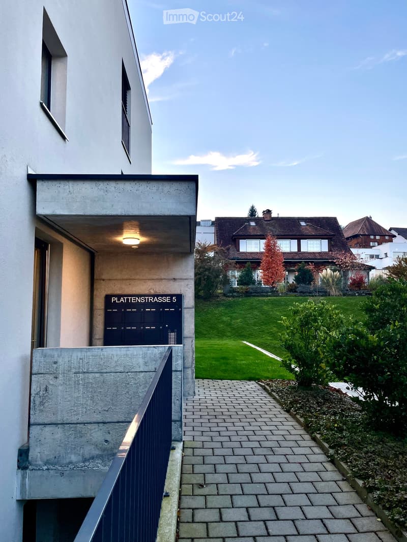 Modern building entrance with a concrete facade, blue mailbox, and a well-maintained garden area with a lawn, pathway, and shrubs.