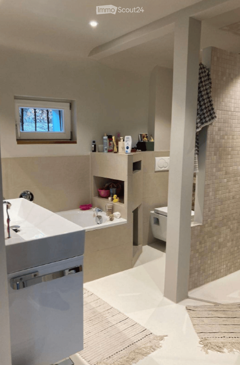 Modern bathroom with a white sink, bathtub, toilet, and shower area. A shelf holds various toiletries and a towel hangs on the wall. The space is well-lit with a window providing natural light.