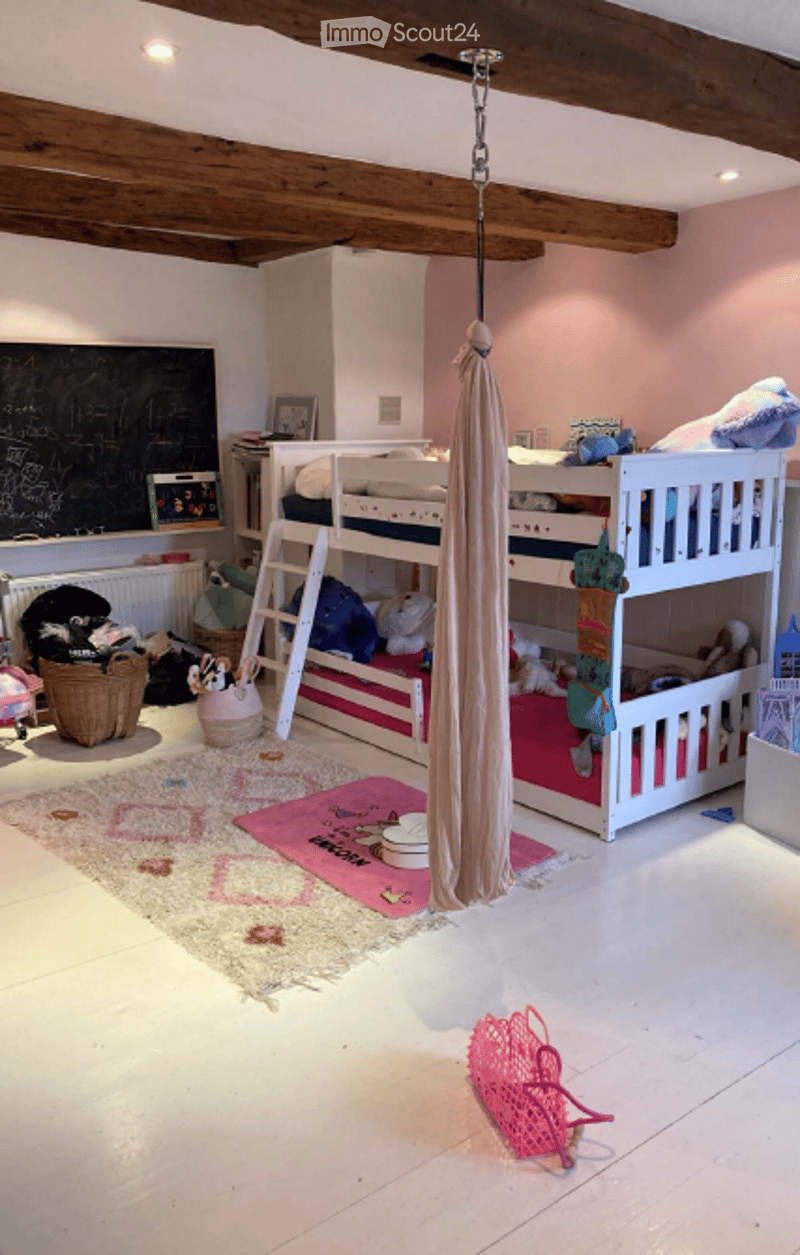A cozy children's bedroom with a loft bed, colorful toys, a hanging fabric swing, and a chalkboard wall. The room features exposed wooden beams, a white tiled floor, and a soft, pink and cream color scheme.