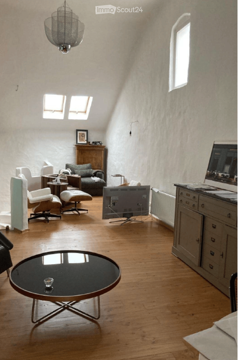 A cozy living room with wooden flooring, a large window, and skylights. Features include a sofa, coffee table, chairs, ottoman, a TV screen, and a cabinet.