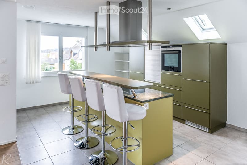 Modern kitchen with a bar counter, four white stools, green cabinets, and an integrated oven. The space features a skylight and large windows offering a view of the neighborhood.