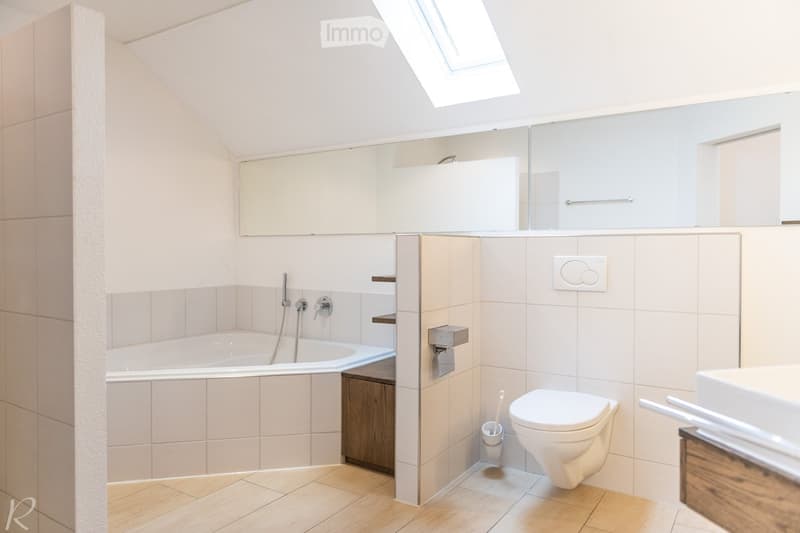 Modern bathroom with a bathtub, toilet, sink, mirror, and built-in cabinet. Light-colored tiles and wooden flooring.