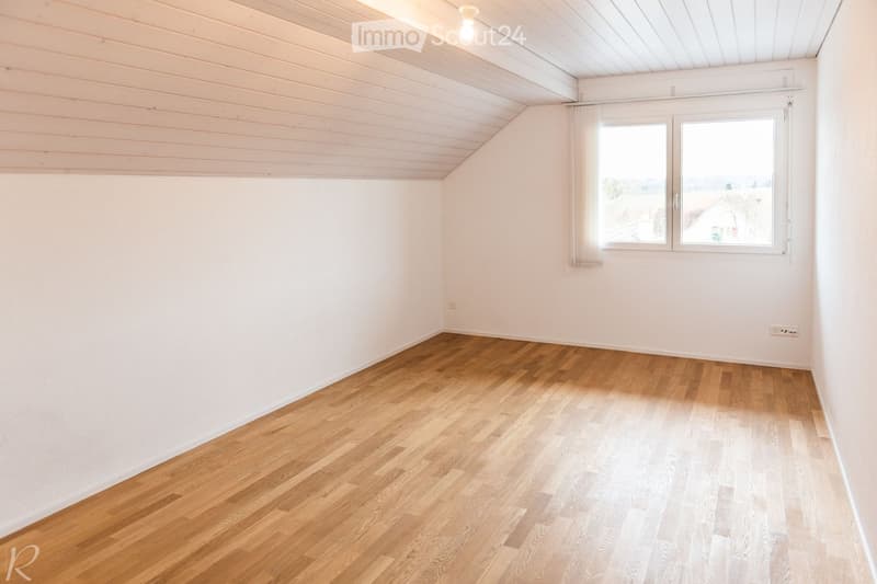 Empty room with light wooden flooring, white walls, and a slanted white ceiling. Two windows with white frames and curtains are visible on one wall.