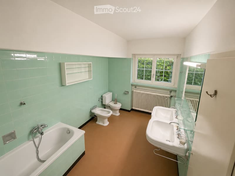 Bathroom with mint green tiles, white fixtures, bathtub, double sink, toilet, bidet, radiator, and a window with a view of greenery.