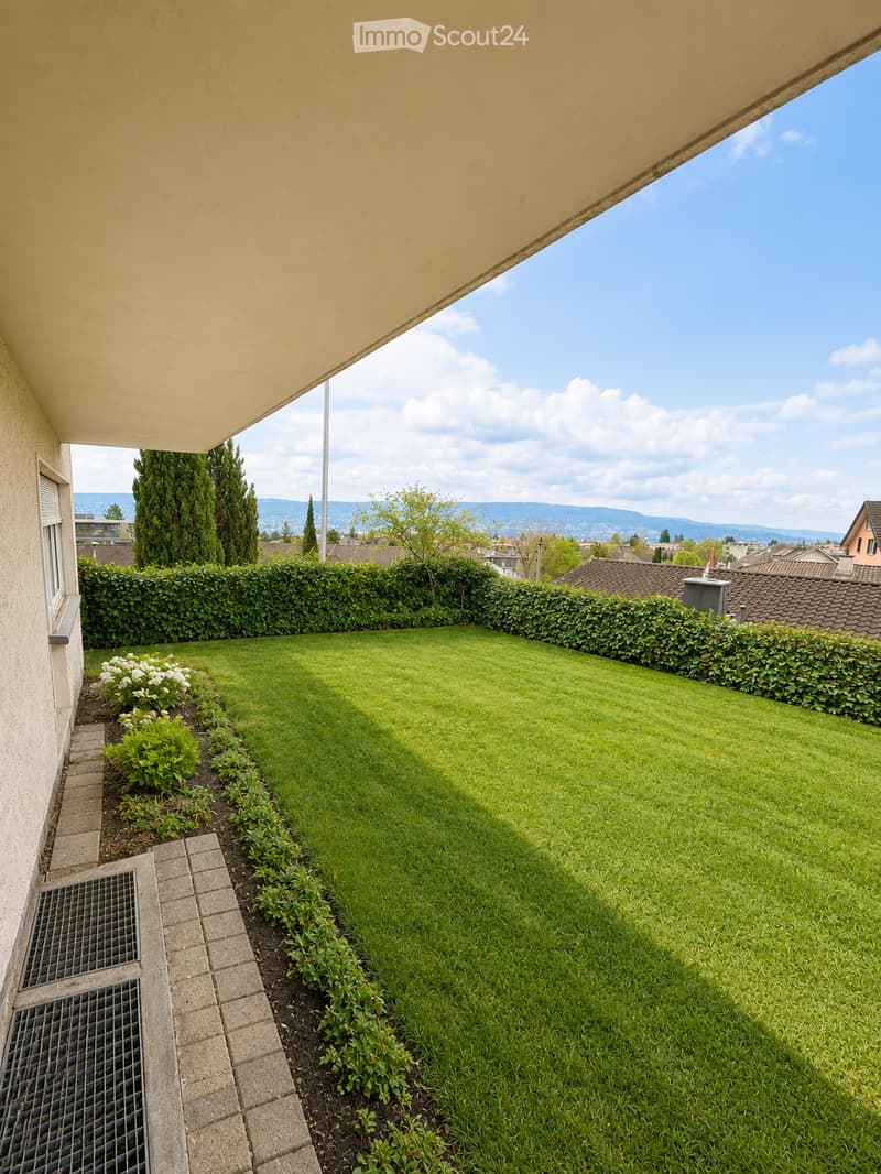 Well-maintained garden with a neatly trimmed lawn, surrounded by hedges and small flower beds, with a clear view of the city and mountains in the background.