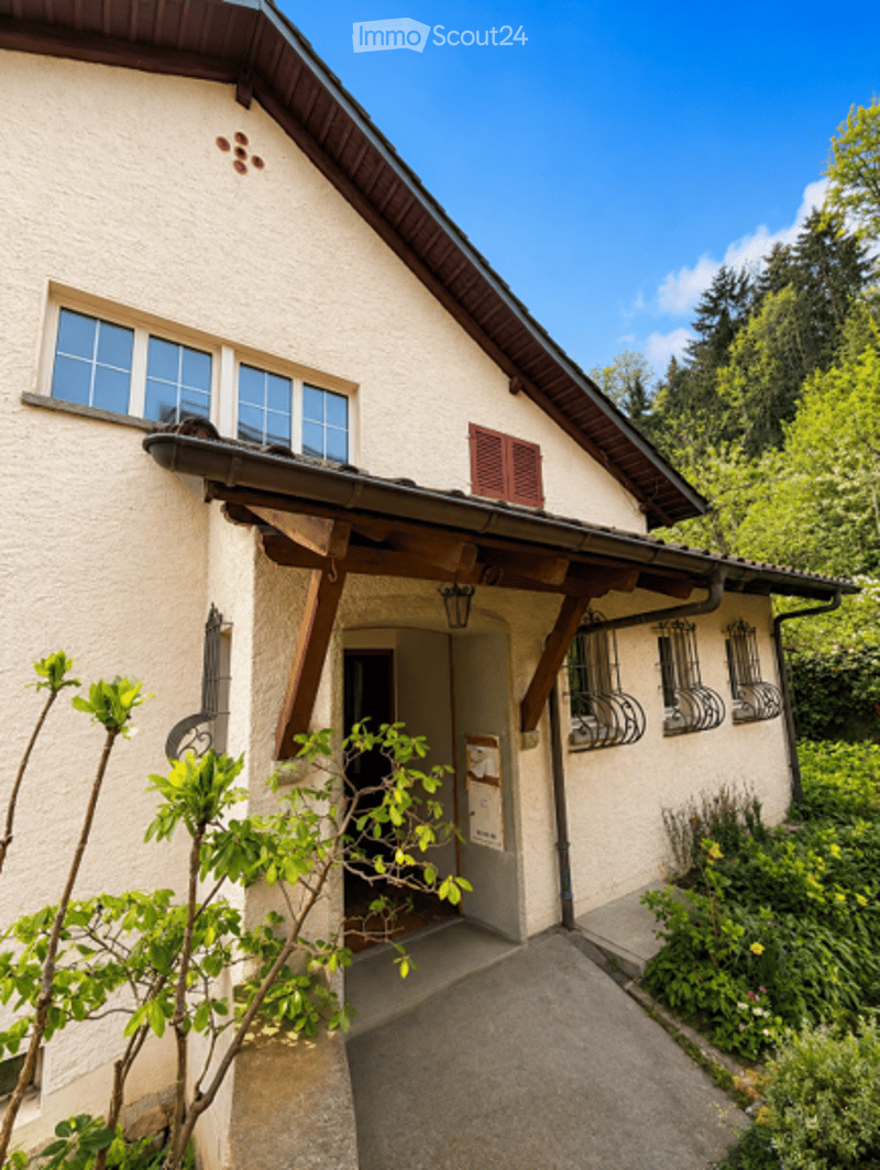 A charming, light-colored house with a wooden entrance canopy, multiple windows, and a small garden area with greenery and flowers.