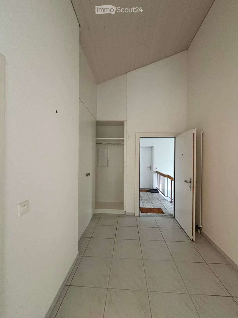 Empty hallway with tiled floor, white walls, and an open door leading to another room with a visible staircase and railing.