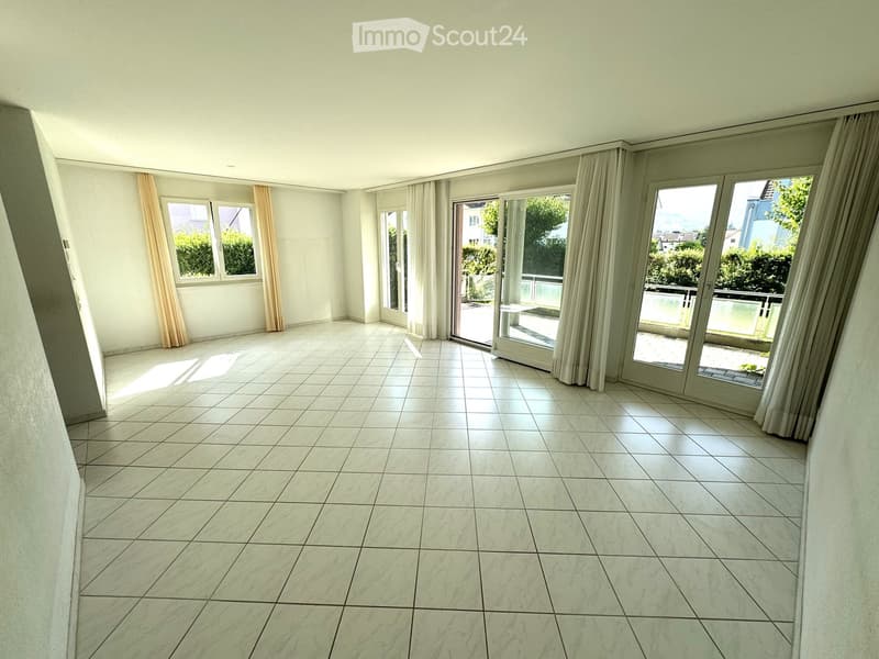 Empty room with white tiled floor, large windows with curtains, and sliding glass doors leading to a balcony.