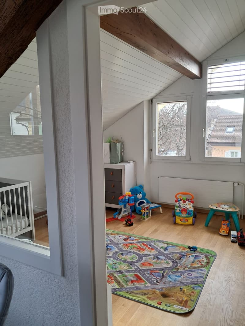 Bright attic room with wooden flooring, slanted ceilings, large windows, crib, toy car, toy truck, toy table, toy chair, toy bear, toy walker, toy play structure, storage cabinet, and handbag.