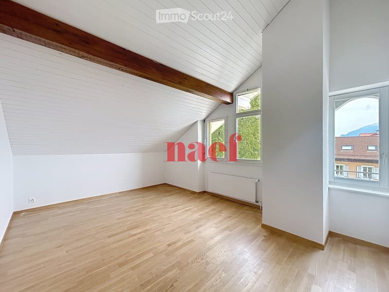 Empty room with wooden flooring, white walls, and large windows offering a view of greenery and a neighboring building.
