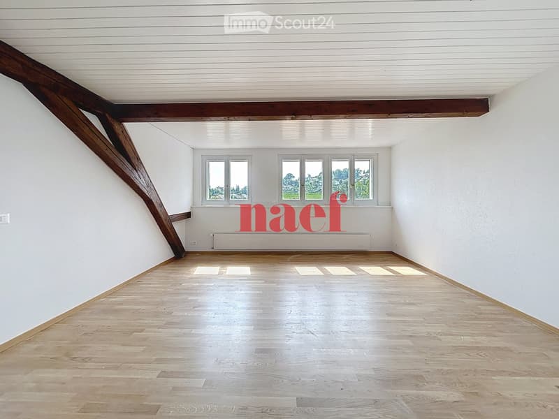 Empty room with wooden flooring, white walls, and large windows, featuring exposed wooden beams on the ceiling.