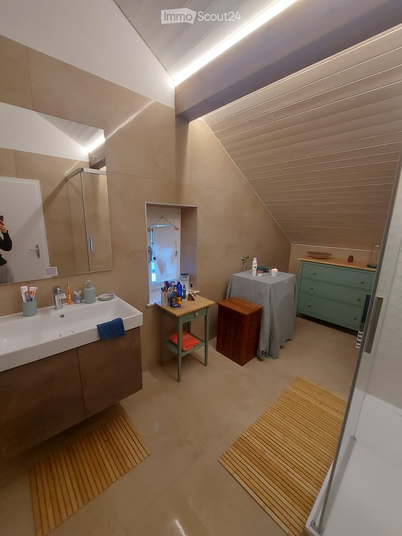 Modern bathroom with a large sink, mirror, and wooden floor. Features include a small table with toiletries, a stool, a covered table, and a green cabinet.
