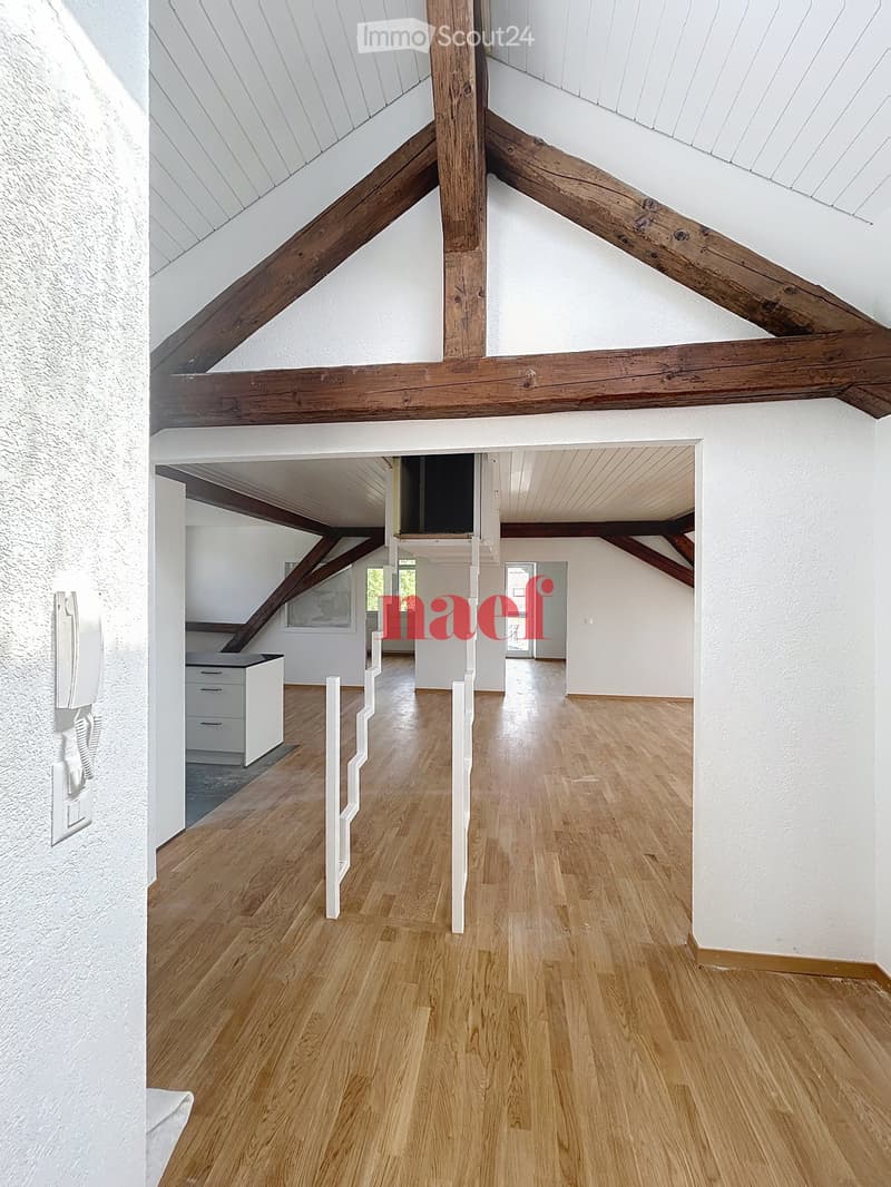 Empty room with wooden flooring, white walls, and exposed wooden beams on the ceiling. The space is open and well-lit with natural light coming from a window in the background.