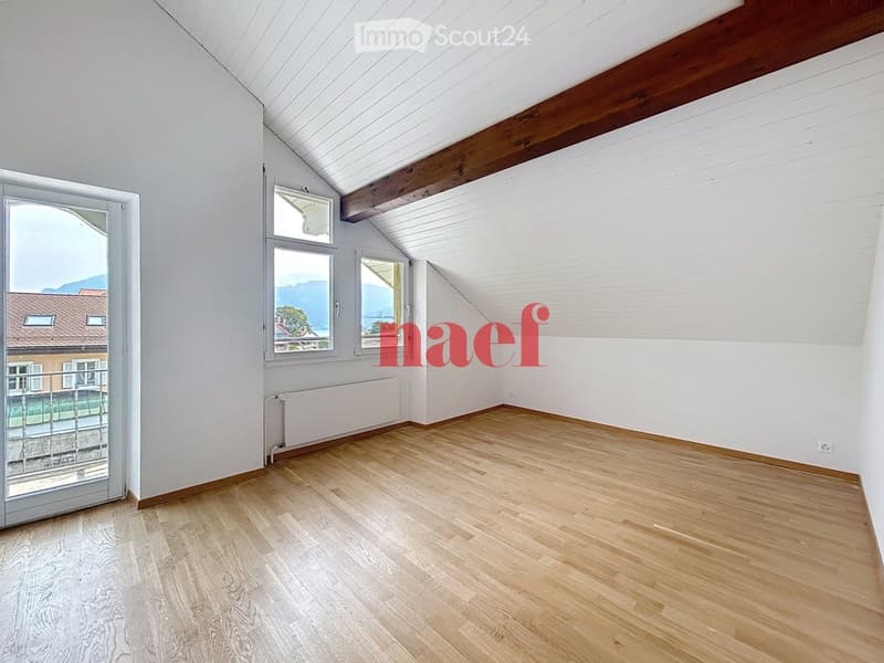 Empty room with wooden flooring, white walls, and large windows offering a scenic view. The room features exposed wooden beams on the ceiling and a radiator along one wall.