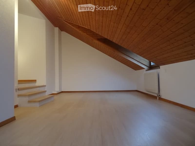 Empty room with wooden flooring, white walls, and a wooden staircase leading up, featuring a slanted wooden ceiling and a small skylight window.